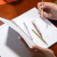 Person holding a pen over an open planner with other pens on the page.