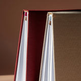 Close-up of a hardcover notebook or journal with a deep burgundy red fabric cover and white pages, standing next to another beige textured cover against a warm brown background.