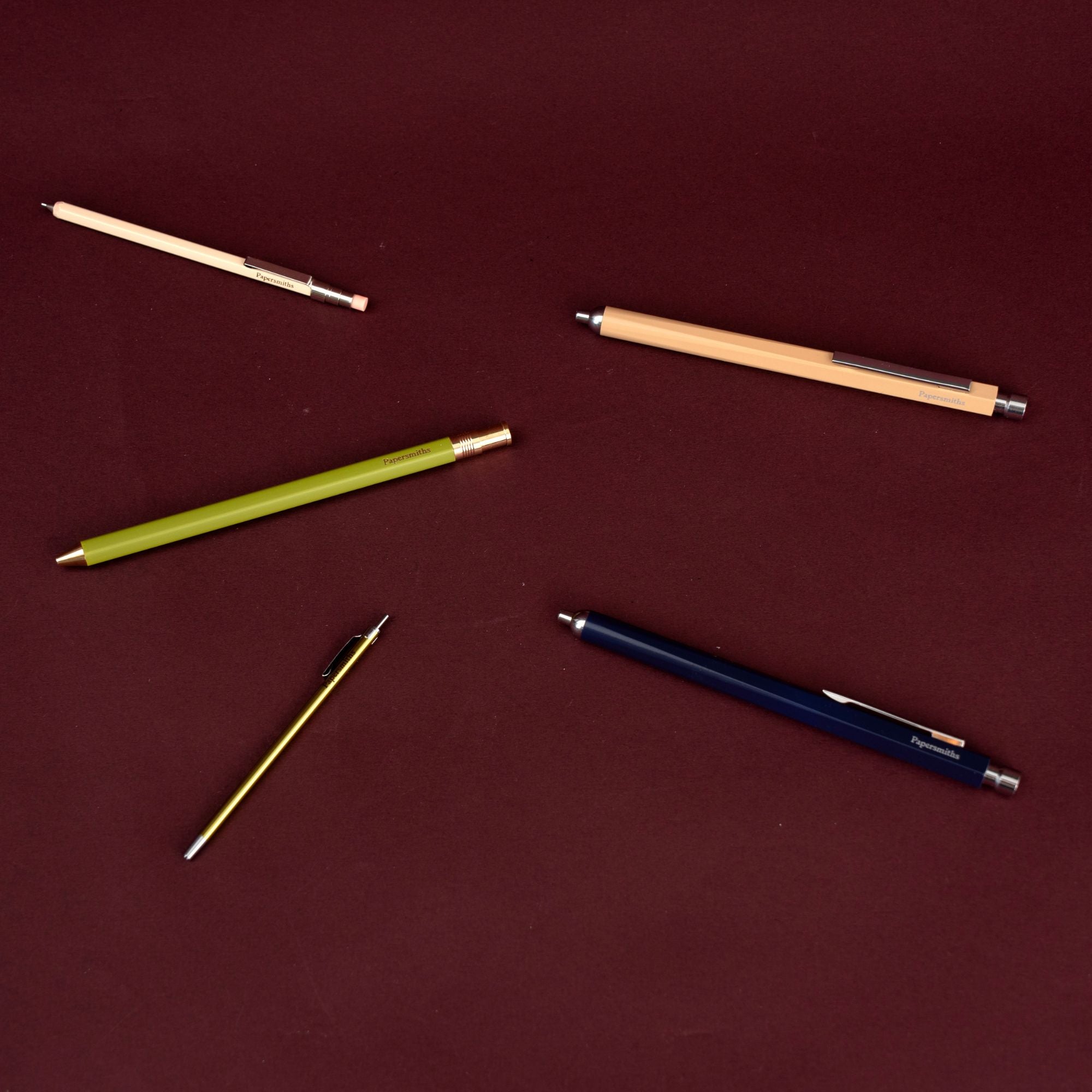 Five retractable mechanical pencils in various colors (beige, light green, navy blue, and silver) arranged on a solid dark red background.