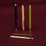 Three retractable ballpoint pens in navy blue, beige, and olive green are displayed upright on a dark maroon background, with two additional pens lying horizontally in front.