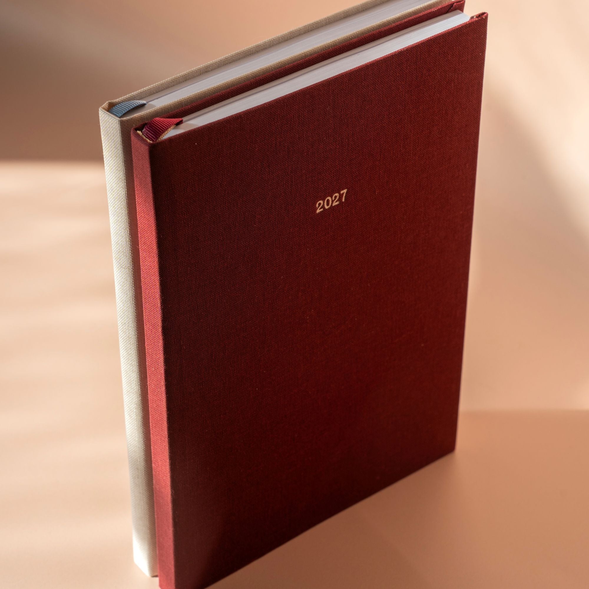A hardcover 2027 journal with a textured deep burgundy cover and gold foil '2027' text, standing upright on a warm beige surface under soft directional sunlight.