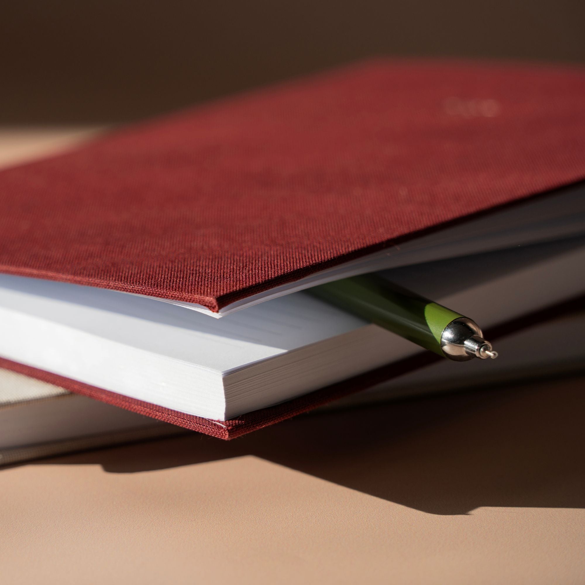 A close-up of a dark red fabric-covered notebook or journal with white pages, partially covered by a matching red cover, with a green pen resting across the edge on a warm beige surface.