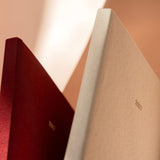 Two hardcover books or notebooks standing upright close-up, one in deep burgundy and one in warm cream, both with small gold '2027' text on the spines, against a softly lit beige background.