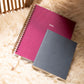 Two spiral-bound planners, one pink and one gray, on a textured surface.