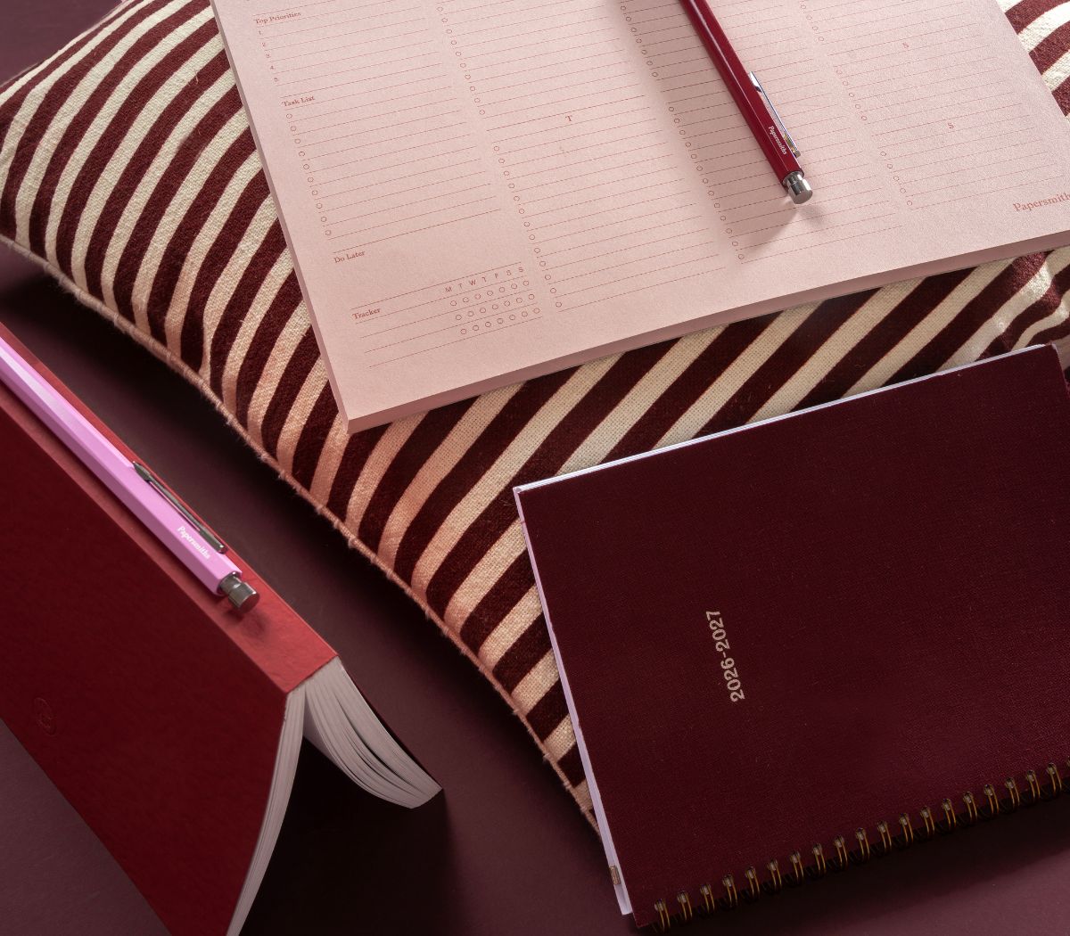 A styled flat-lay of stationery items on a dark surface, including a striped brown and cream pillow, a pink pen, an open lined notebook, a closed burgundy notebook with gold spiral binding, and another closed burgundy notebook.