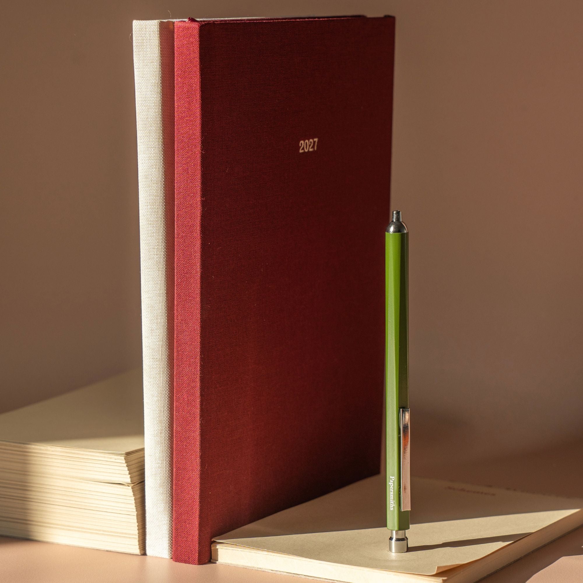 A dark red fabric-covered 2027 planner or journal stands upright on a beige surface, next to a green pen resting on a light notebook. The scene is lit by warm directional sunlight, casting soft shadows against a neutral beige background.