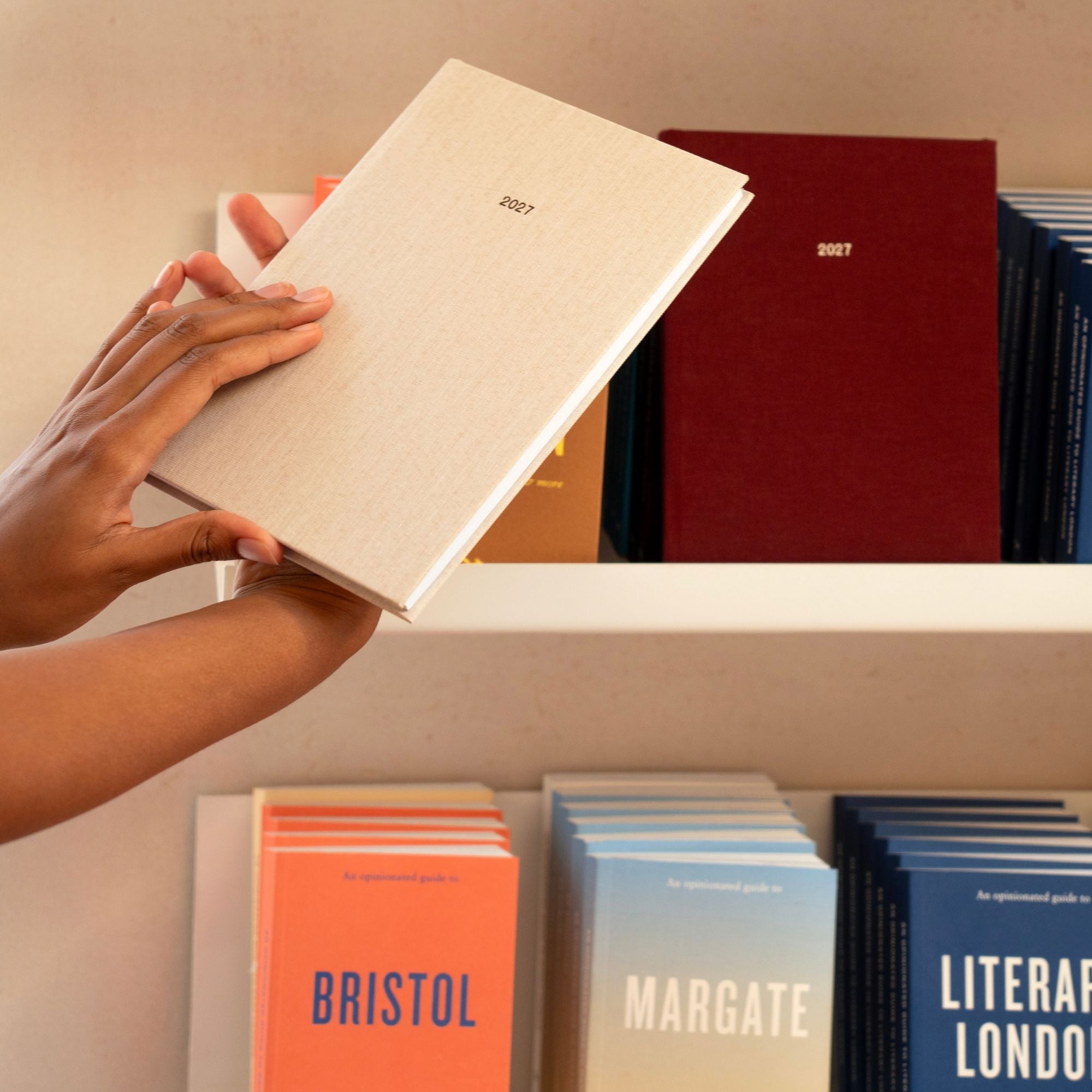A person's hands holding a light beige 2027 edition of Papersmiths 2027 diary against a beige bookshelf, with other colorful guidebooks in the background.