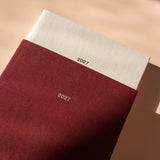 A close-up of a 2027 fabric-covered journal or planner, showing a deep burgundy cover with a white top flap and the year '2027' printed in gold. The photo is styled with warm, directional sunlight and soft shadows on a beige surface.