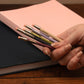 Hand holding a set of colorful pens with a pink notebook in the background