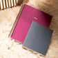 spiral-bound planners, one maroon  and one gray, on a textured surface.