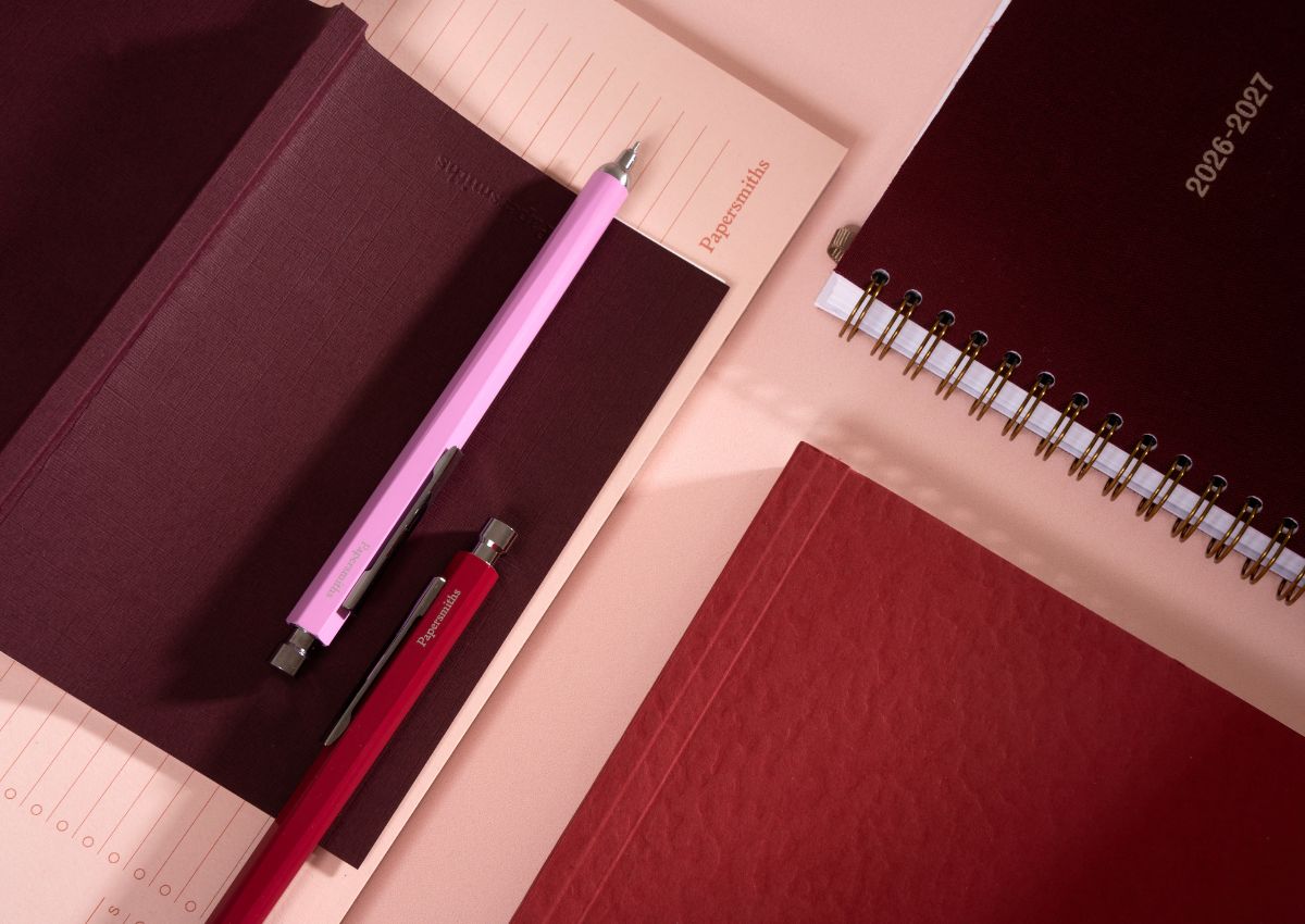A styled flat-lay of stationery items on a peach background, including a dark burgundy planner or notebook, a pink pen, a red pen, and a spiral-bound planner.