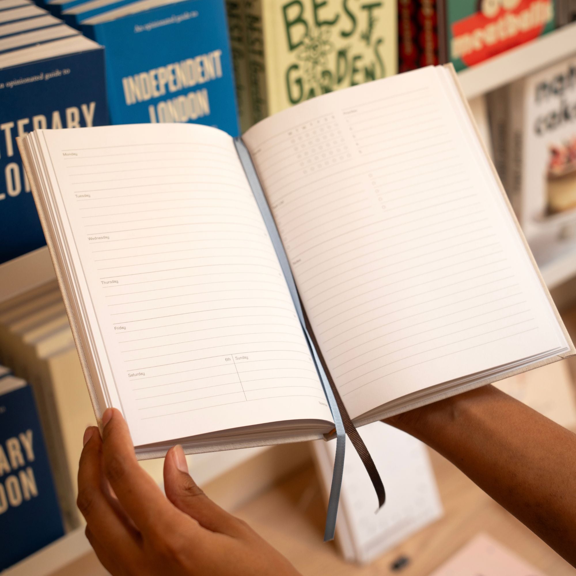 A person holding an open lined journal or planner with a blue ribbon bookmark, photographed in a real bookstore environment with bookshelves and other books blurred in the background.