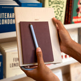 A person holding a stack of books in a library or bookstore, featuring a beige hardcover book with '2027' printed on it, a dark brown planner or notebook, and a light blue pen.