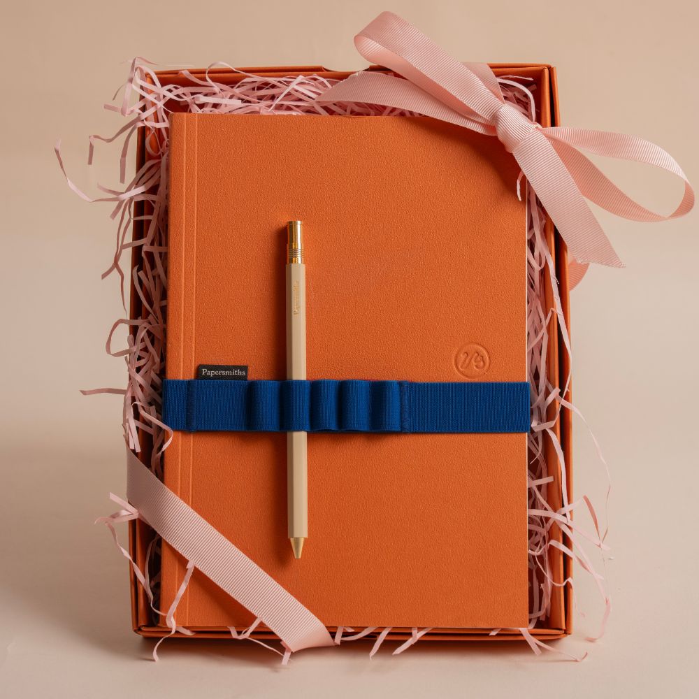 Orange notebook with a pen and decorative ribbon in a gift box on a beige background