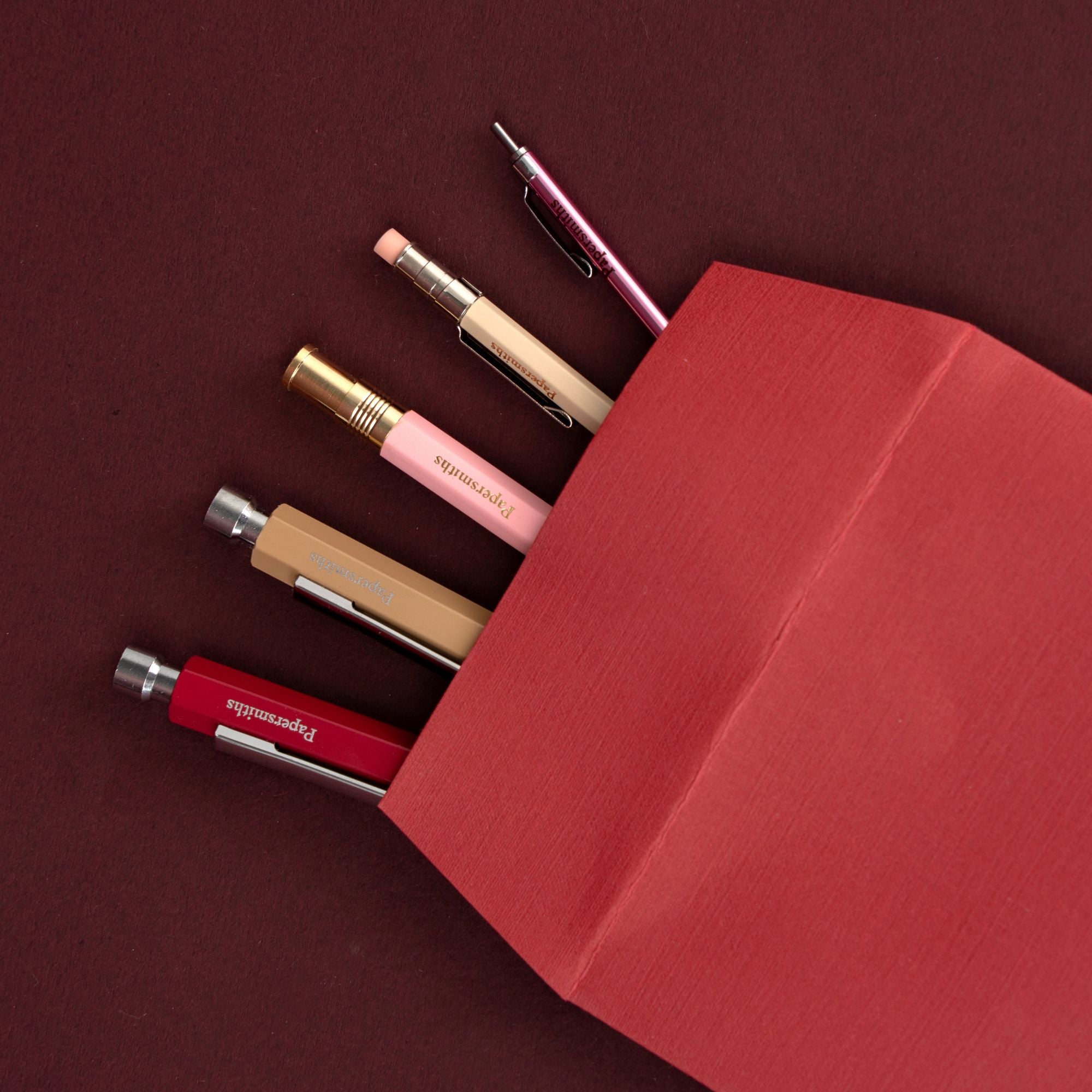 A red textured paper envelope or pouch on a dark red background, with several pens and a pencil partially sticking out from the opening. Visible pen brands include Papersmiths
