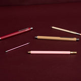 Four slim retractable pens in red, beige, pink, and metallic rose gold, arranged diagonally on a dark burgundy background.