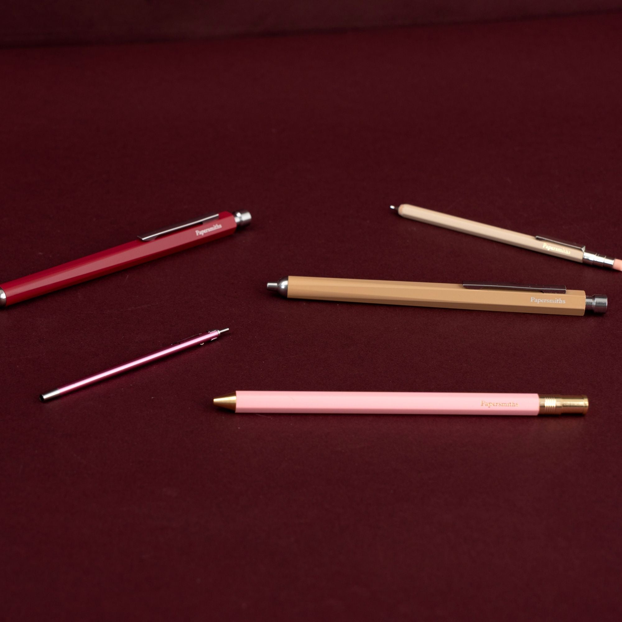 Four slim retractable pens in red, beige, pink, and metallic rose gold, arranged diagonally on a dark burgundy background.
