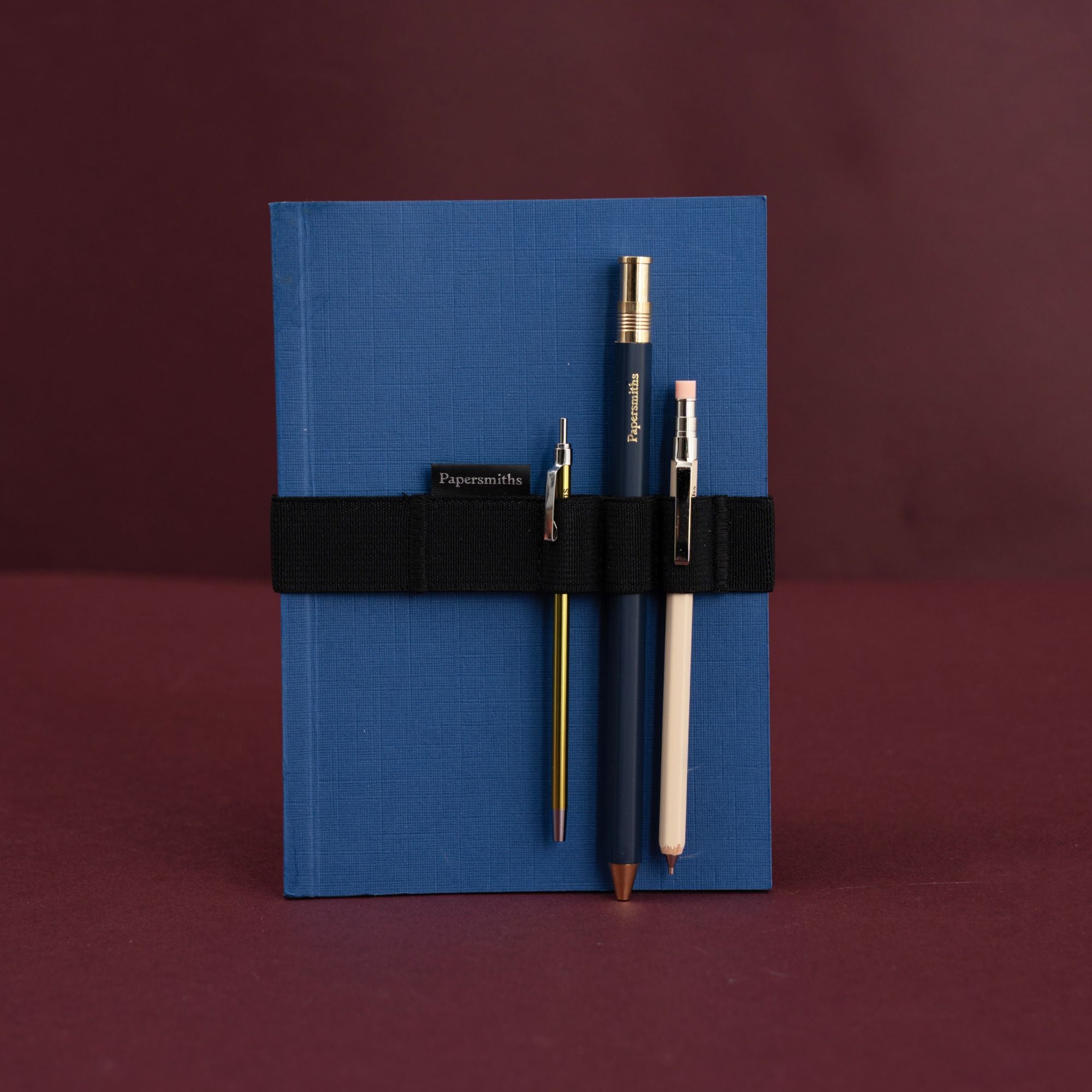 A blue softcover notebook with a black elastic band holding three pens, photographed against a dark burgundy background. The brand name 'Papersmiths' is visible on the pen labels.