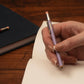 Hand holding a purple pocket pencil over a notebook on a wooden surface