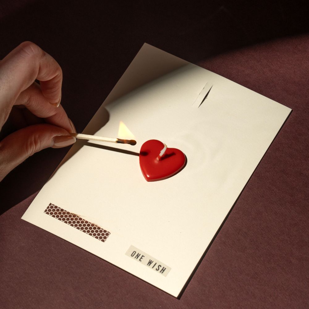 Red heart-shaped candle being lit with a match on a card labeled 'One Wish'.