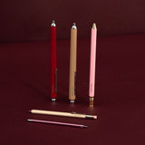 Three slim retractable ballpoint pens in red, tan, and pink are displayed upright on a dark burgundy background, with one tan pencil and a pink pocket pen lying horizontally in front.