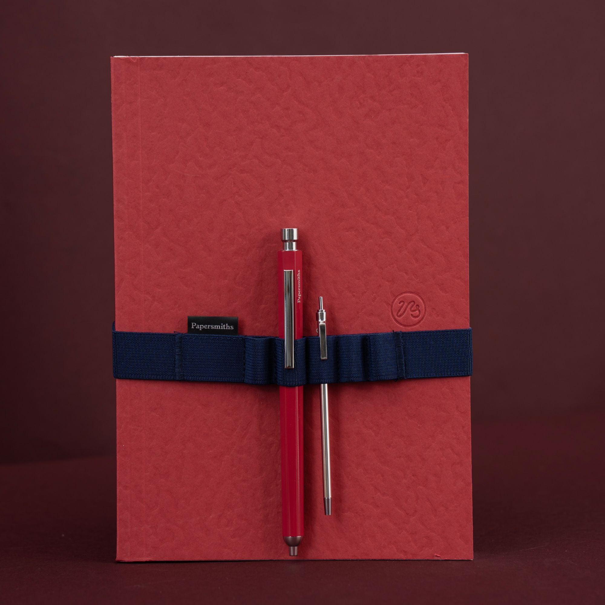 A red textured notebook or journal with a navy blue elastic band holding two pens, photographed against a dark reddish-brown background. The band has a small black label reading Papersmiths and a small embossed logo on the right side.