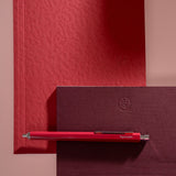 A red papersmiths pen with silver accents resting horizontally on a dark burgundy textured notebook, with a matching red textured notebook in the background against a dusty pink wall.