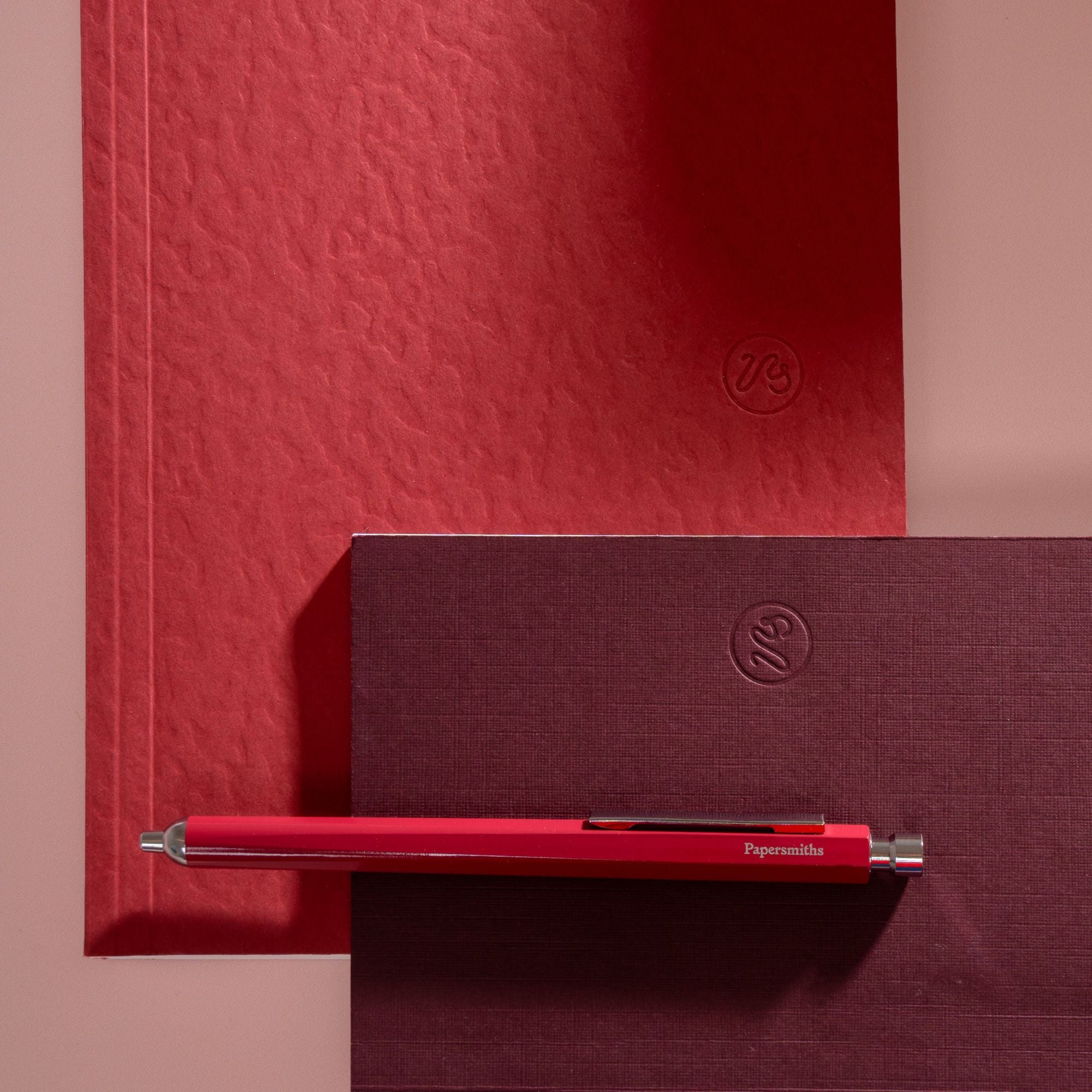 A red papersmiths pen with silver accents resting horizontally on a dark burgundy textured notebook, with a matching red textured notebook in the background against a dusty pink wall.