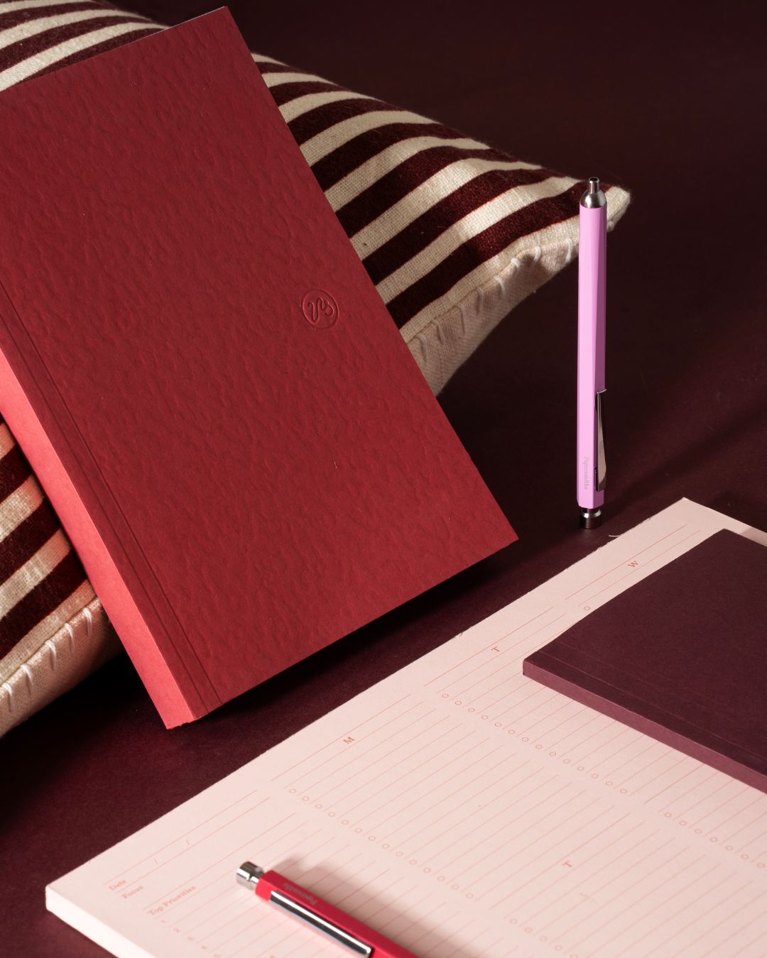 A dark red textured hardcover notebook or journal standing upright on a striped cushion, with a pink pen beside it and a white lined notepad with a matching dark red pad and red pen in the foreground.