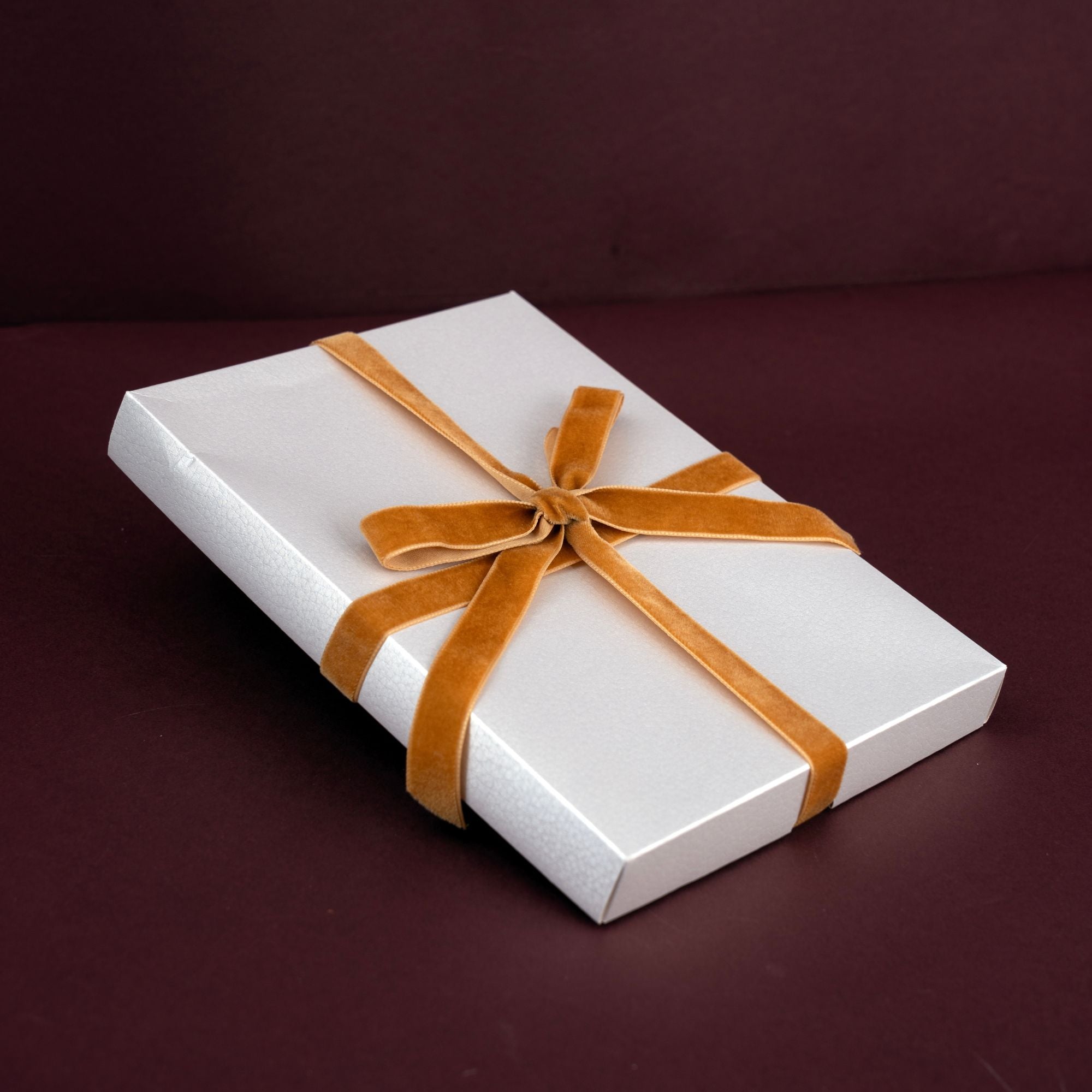A white rectangular gift box wrapped with a wide tan ribbon tied in a bow, photographed on a dark burgundy background.